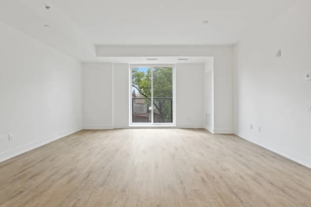 Ein leerer Raum mit weißen Wänden, einem großen Fenster und einem hellen Holzfußboden. Das Fenster bietet einen Blick auf Bäume und ein Gebäude.