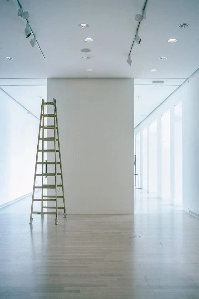 Eine Leiter steht in einem minimalistischen Museumsraum vor einer weißen Wand. Der Raum ist hell und leer, mit einem glänzenden Holzboden.