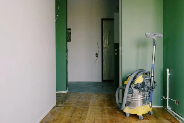 Ein gelber Staubsauger steht in einem Zimmer mit frisch gestrichenen Wänden in Weiß und Grün. Das Zimmer befindet sich in einer Renovierungsphase, was durch freiliegende Kabel und den unfertigen Bodenbelag deutlich wird.