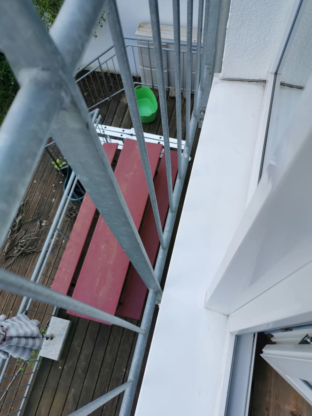 Ein Blick von oben auf einen Balkon mit einem grauen Geländer, einem roten Tisch und einem grünen Eimer. Die Perspektive zeigt auch einen Teil der Hauswand und ein geöffnetes Fenster.