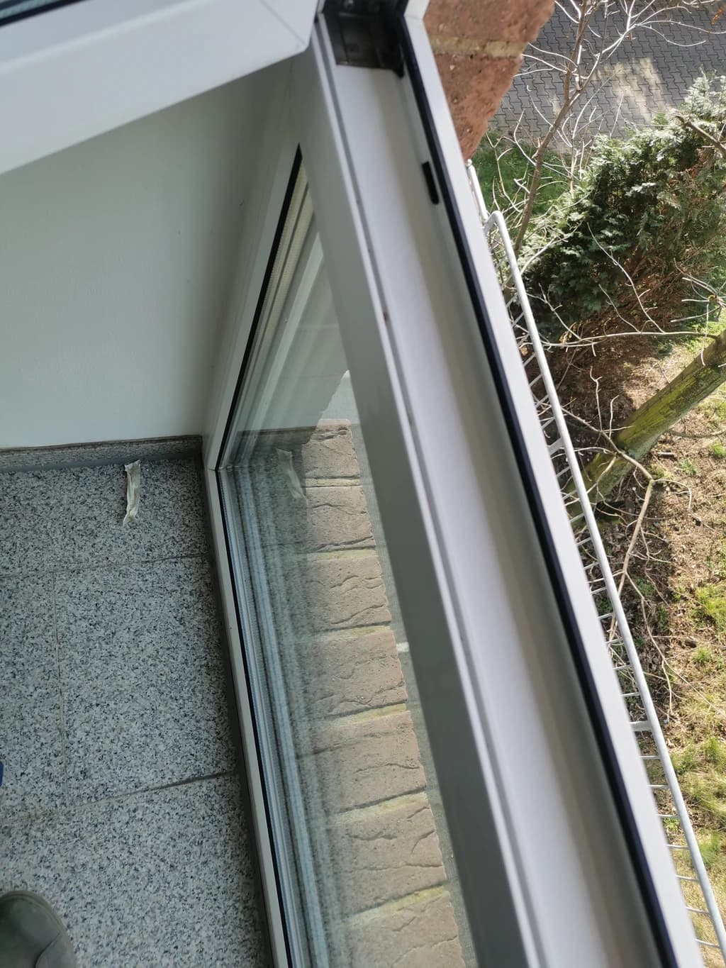 Ein Blick aus einem geöffneten Fenster auf einen Balkon mit Blick auf einen Garten oder Aussenbereich. Man sieht einen Teil des Fensters, den Boden des Balkons und im Hintergrund Bäume und Pflanzen.