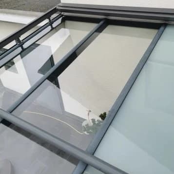 Nahaufnahme eines Glasdachs über einer Terrasse mit Metallrahmen. Das Glasdach reflektiert die Umgebung.