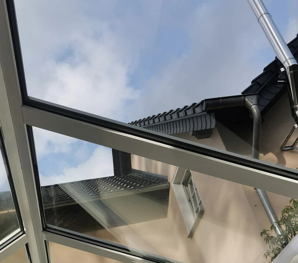 Eine ungewöhnliche Perspektive auf ein Hausdach und den Himmel durch einen Fensterrahmen. Das Bild zeigt das Dach, den Himmel und Teile der Hausfassade, die durch das Glas des Fensters zu sehen sind.