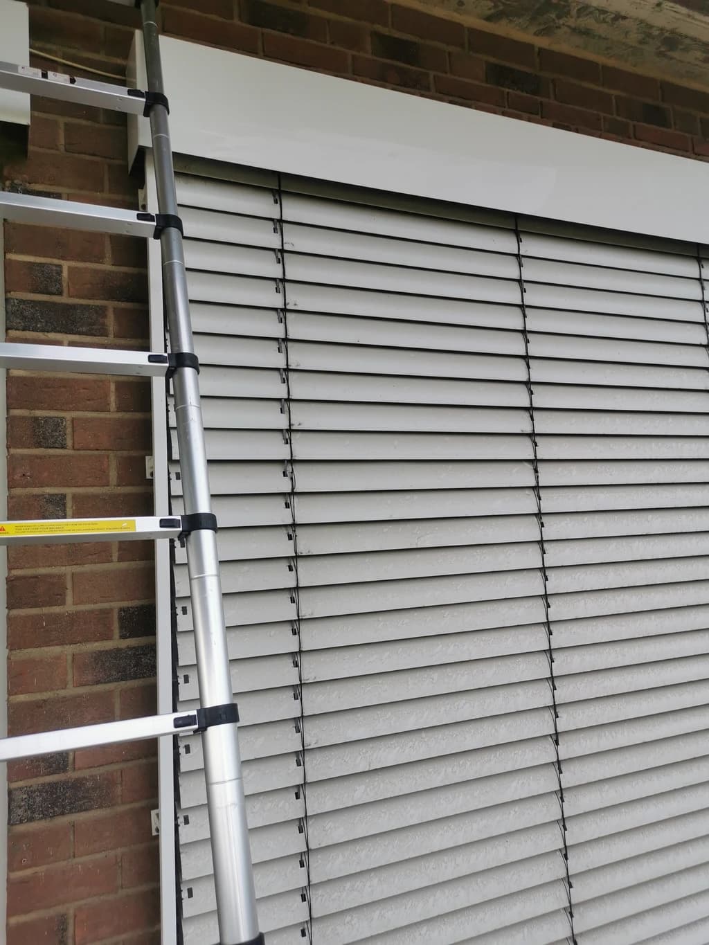 Eine Leiter steht an einer Hauswand mit geschlossenen Jalousien vor einem Fenster. Die Jalousien sind grau und die Hauswand ist aus roten Ziegelsteinen.