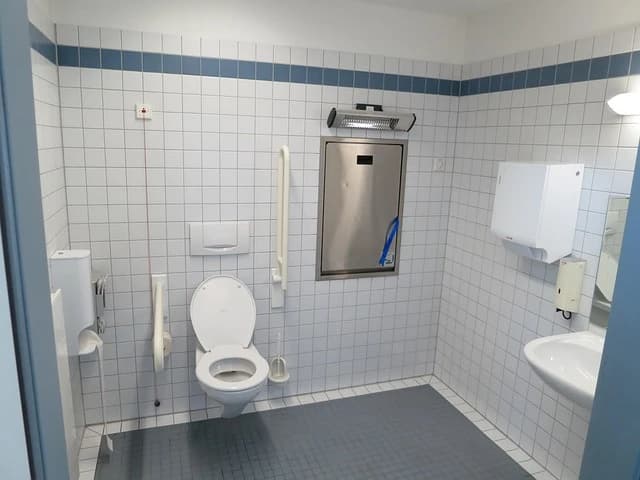 Ein barrierefreies, öffentliches Behinderten-WC mit Toilette, Waschbecken, Wickeltisch und Haltegriffen. Der Raum ist hell gefliest und mit blauen Akzenten versehen.