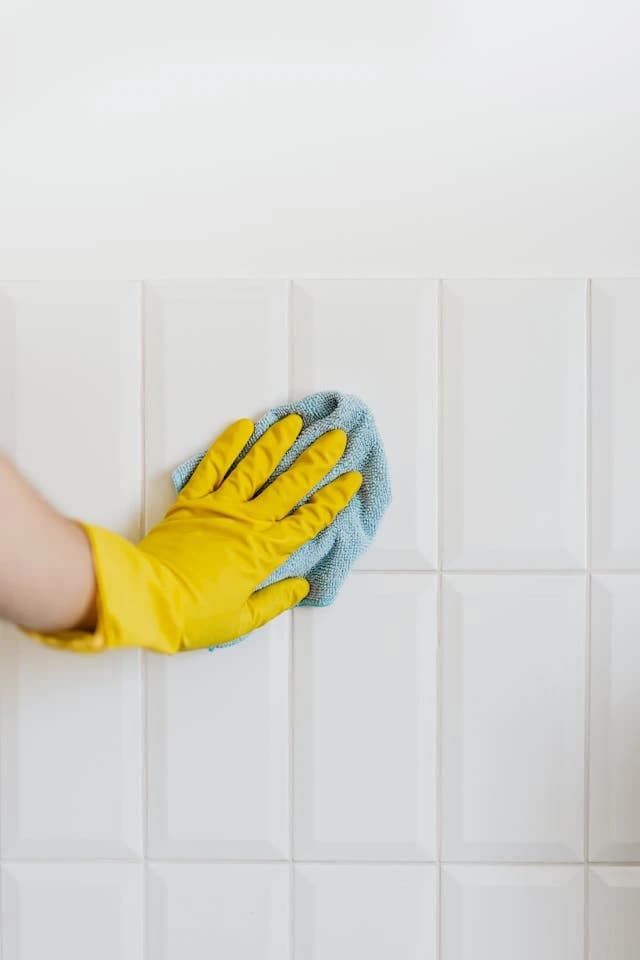 Eine Hand mit gelbem Gummihandschuh reinigt weiße Fliesen mit einem blauen Lappen. Die Fliesen sind rechteckig und glänzend.
