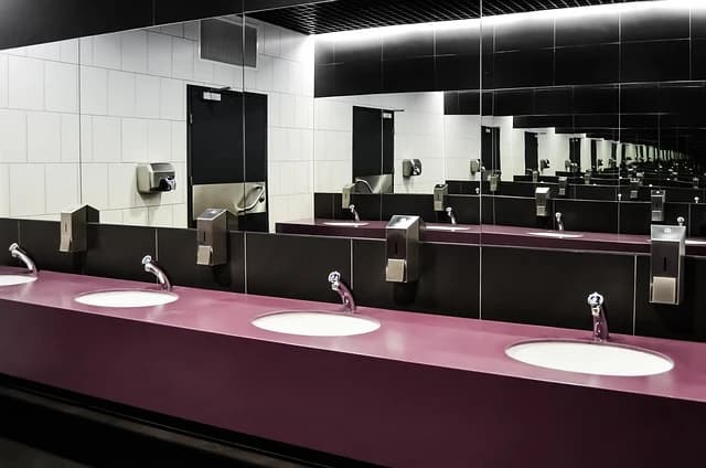 Ein öffentlicher Toilettenraum mit einer Reihe von Waschbecken mit violetten Arbeitsplatten und Spiegeln, die sich unendlich wiederholen. Die Wände sind mit weißen und schwarzen Fliesen bedeckt.