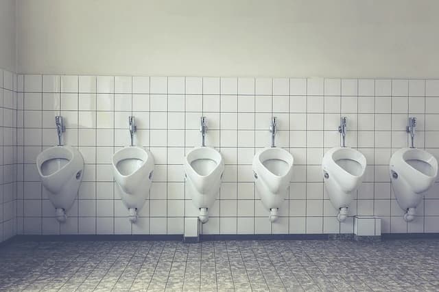 Eine Reihe von sechs weißen Urinalen hängt an einer gefliesten Wand in einer Herrentoilette. Der Boden ist ebenfalls gefliest.