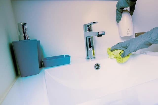Eine Person mit Handschuhen reinigt ein weißes Waschbecken mit einem gelben Tuch. Auf dem Waschbecken befinden sich eine Seifenspender, eine Seifenschale und ein Wasserhahn.