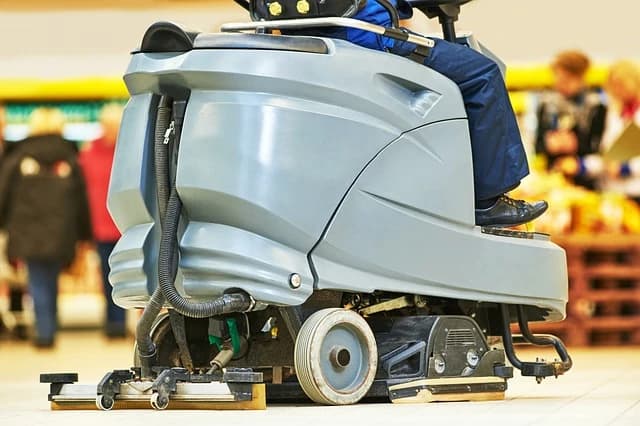 Eine graue Bodenreinigungsmaschine wird von einer Person in einem Supermarkt bedient. Die Maschine reinigt den Boden, während im Hintergrund unscharfe Kunden zu sehen sind.
