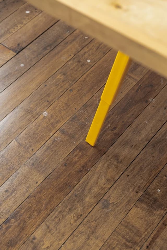 Nahaufnahme eines gelben Tischbeins auf einem Holzboden. Der Holzboden besteht aus einzelnen Brettern.