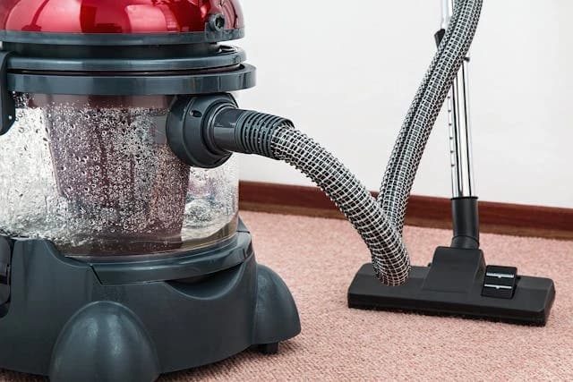 Ein roter Staubsauger steht auf einem Teppich und saugt ihn. Der Staubsauger hat einen durchsichtigen Behälter, in dem man das Schmutzwasser sehen kann.