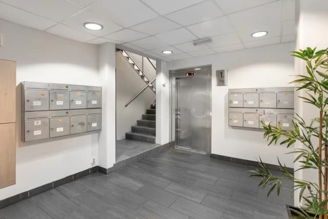 Innenaufnahme eines Treppenhauses mit Aufzug und Briefkästen an den Wänden. Die Böden sind grau gefliest und es gibt eine Treppe im Hintergrund.