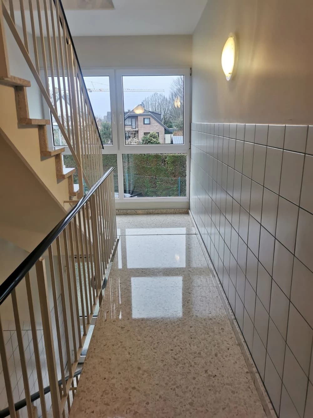 Ein Treppenhaus mit einem großen Fenster, das einen Blick auf ein Wohngebiet bietet. Die Wand ist mit weißen Fliesen verkleidet, und ein Treppengeländer ist im Vordergrund zu sehen.