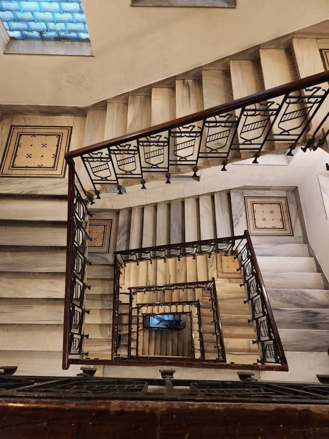 Eine spiralförmige Wendeltreppe aus Marmor mit einem verzierten Geländer aus dunklem Holz und Metall, aufgenommen von oben. Das Treppenhaus ist hell und wird durch ein Oberlicht beleuchtet.