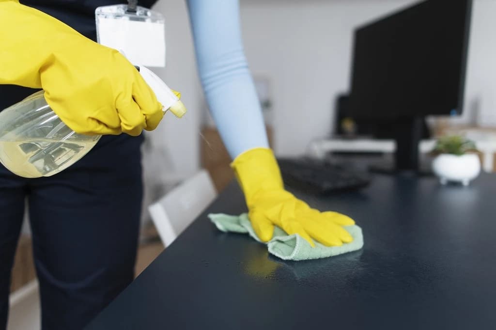 Eine Reinigungskraft mit gelben Handschuhen spritzt Reinigungsmittel auf einen Tisch und wischt ihn mit einem grünen Tuch ab. Im Hintergrund sind ein Computer und eine Pflanze zu sehen.